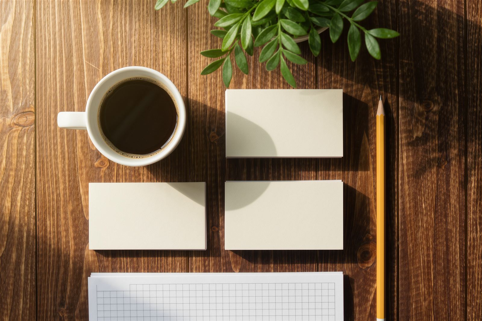 A flat lay of blank place cards, a coffee cup, a pencil and a plain grid planner on a wooden table
