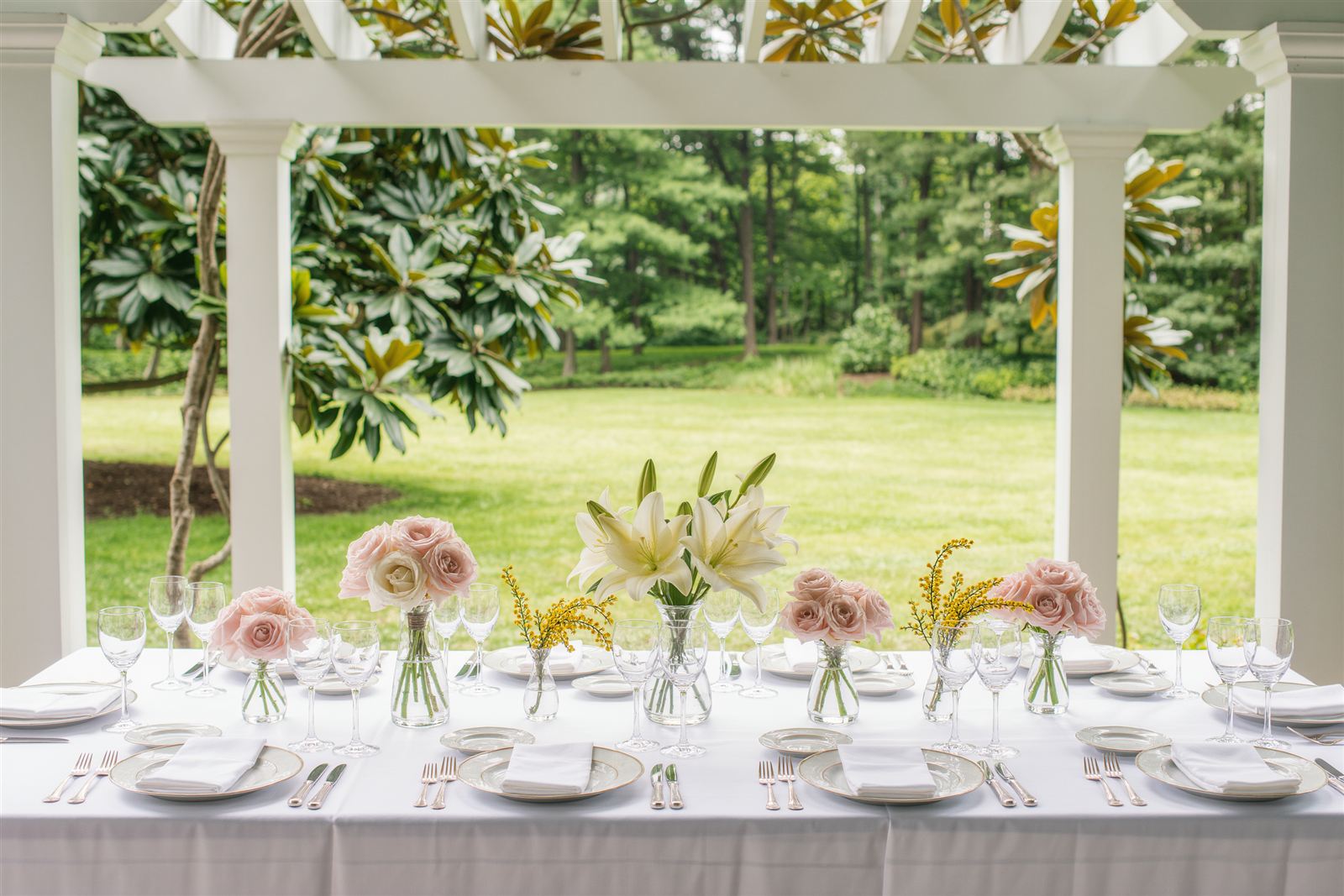 A garden party table under a white pergola set with flowers in glass vases and place settings