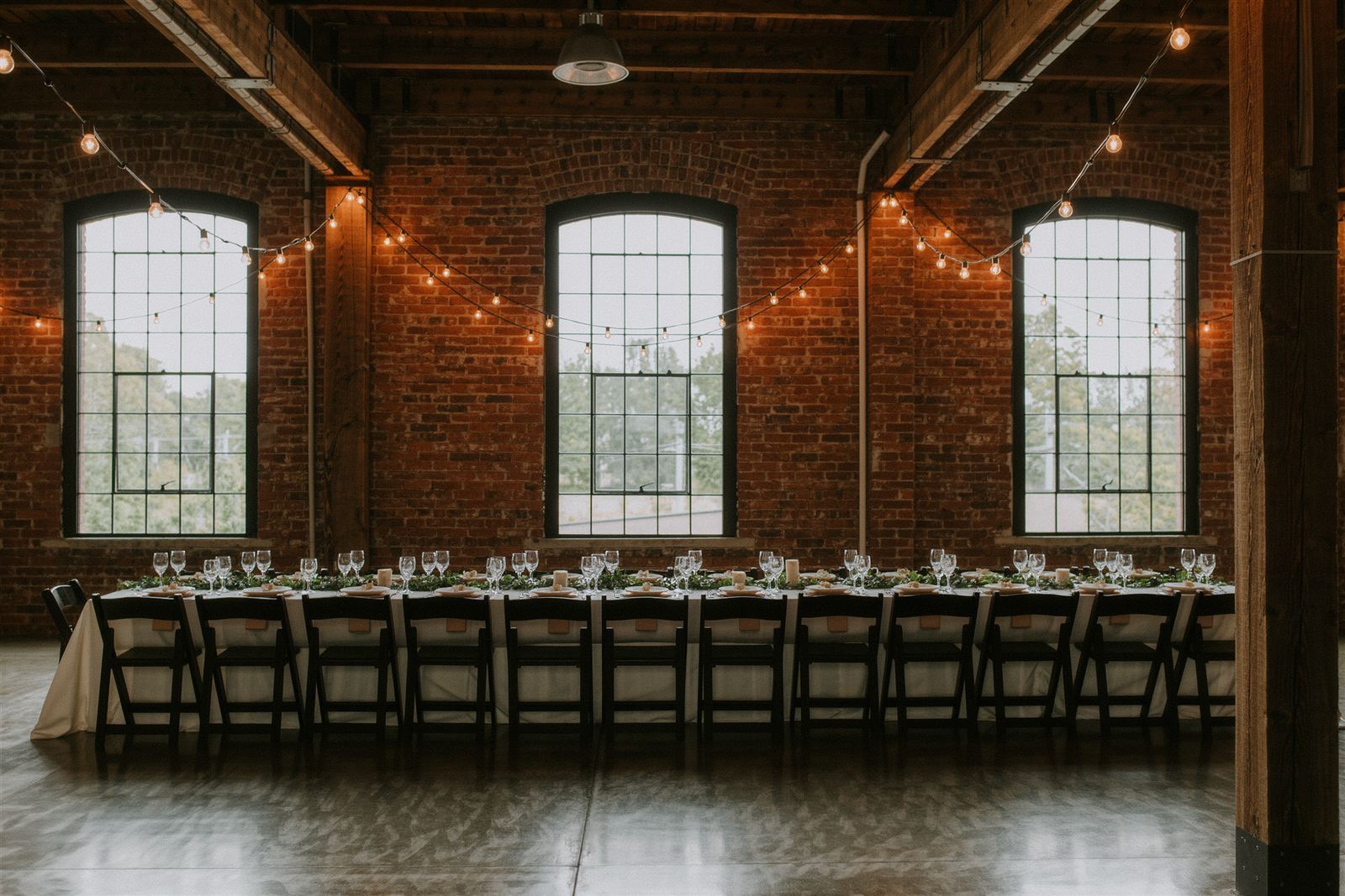 A long banquet table set with white plates and low greenery under strings of warm lights in a brick hall with arched windows