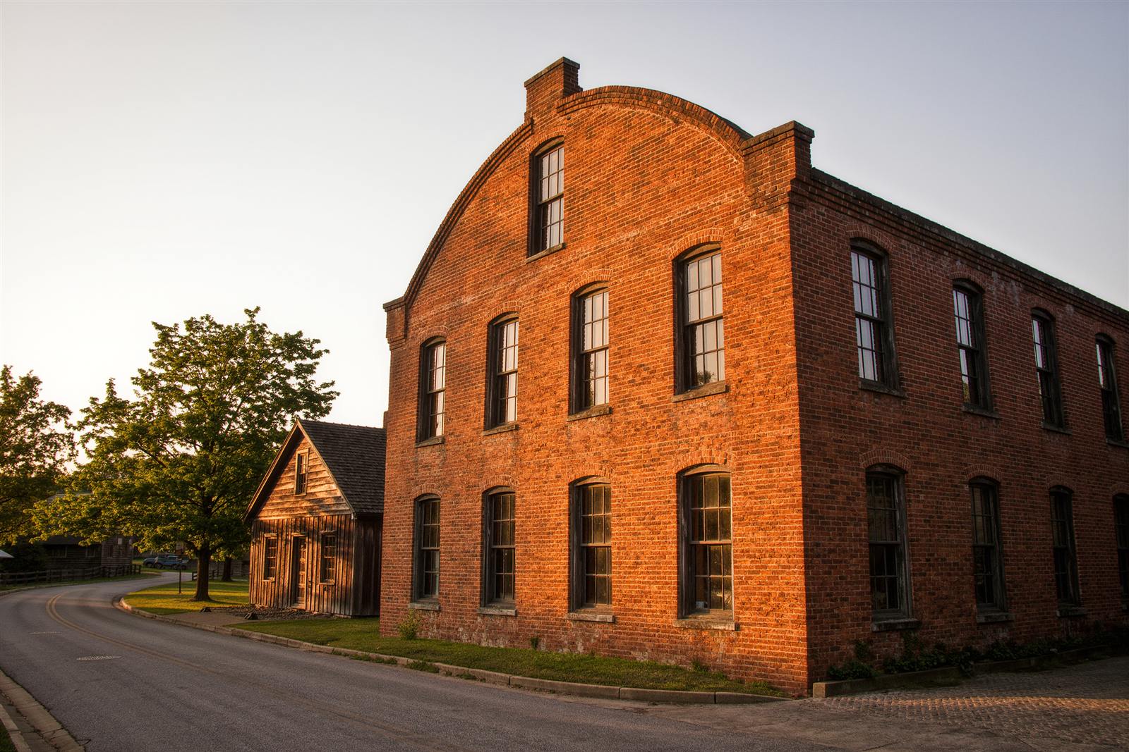 A brick mill building with arched windows beside a quiet street and a wooden mill-village cottage