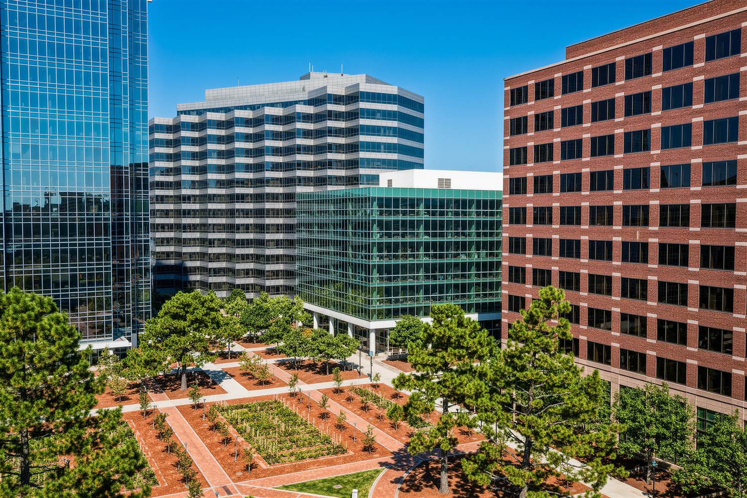 Mid-rise brick and glass office buildings around a landscaped brick plaza with pine and deciduous trees and beds of red clay mulch