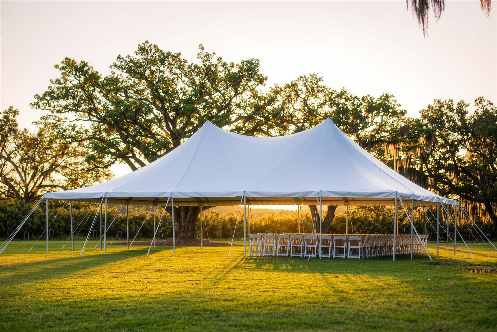 A white open-sided event tent on a green lawn at golden hour with stacked chairs beside it