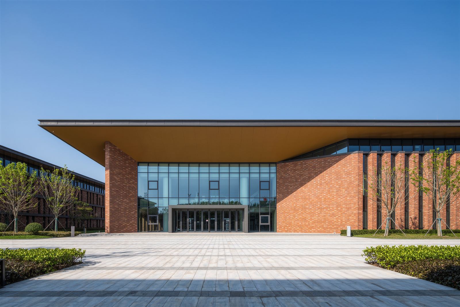 A brick meeting hall with glass doors on a campus plaza with young trees and landscaped beds