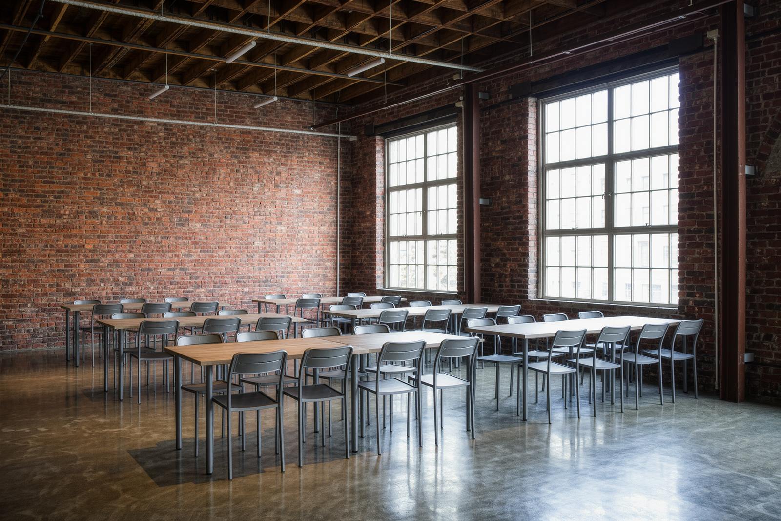 A loft event space with exposed brick, steel-framed windows and empty bistro tables on a concrete floor